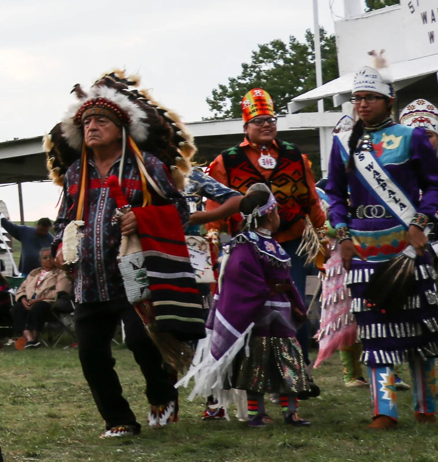 An elder in a full eagle-feather headdress and patterned ribbon shirt dances alongside girls in jingle dresses, one wearing a Miss Wakpala sash.