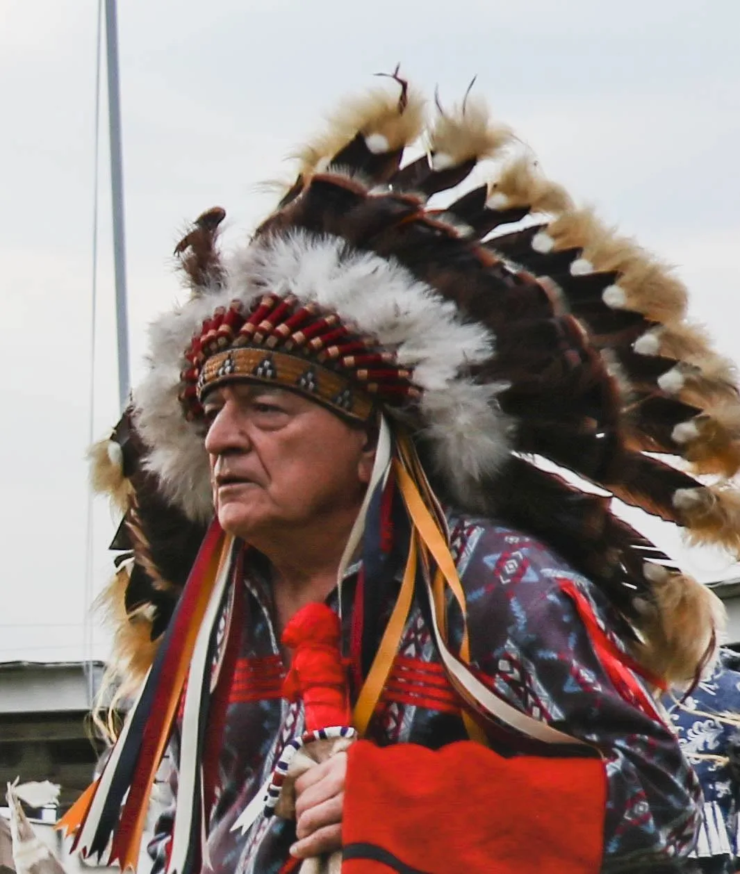 Close-up of an elder in a full feather headdress with colorful ribbons, wearing a patterned shirt and holding a red blanket.