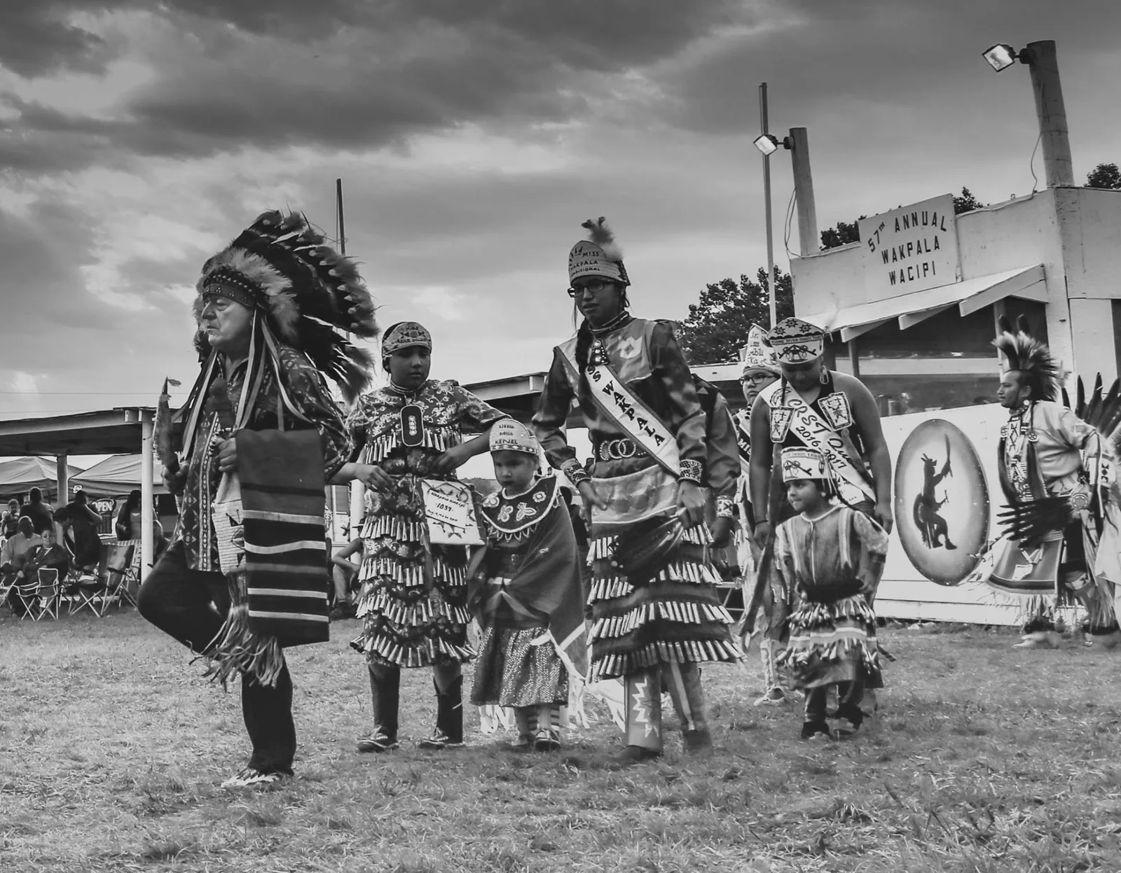 Black-and-white view of an elder in a headdress leading young dancers and royalty in jingle dresses past a stand reading 57th Annual Wakpala Wacipi.