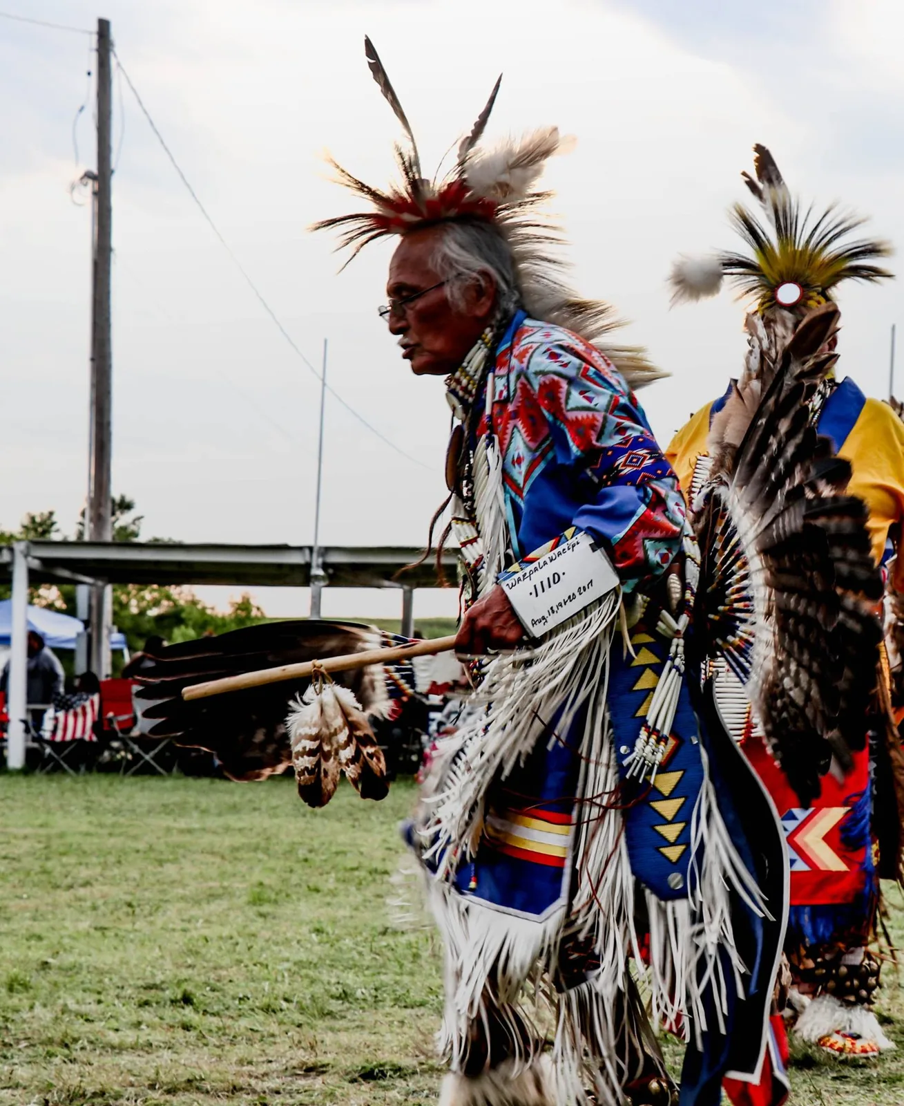An elder dancer with a porcupine roach, glasses and fringed blue regalia carries an eagle feather fan, wearing a Wakpala Wacipi contest number.