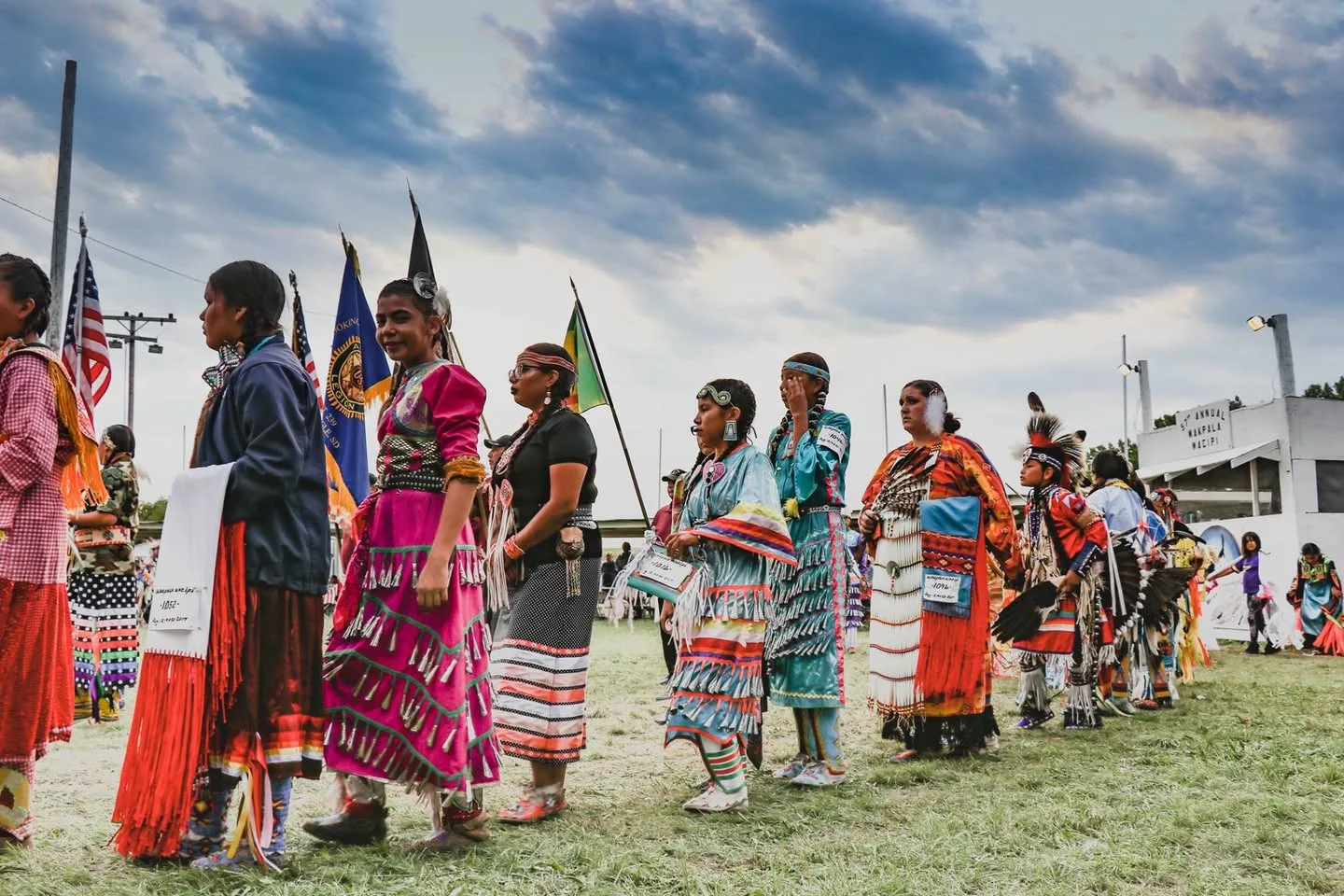 A line of women and girls in colorful jingle and traditional dresses stands on the powwow grounds beneath flags and a cloudy sky.