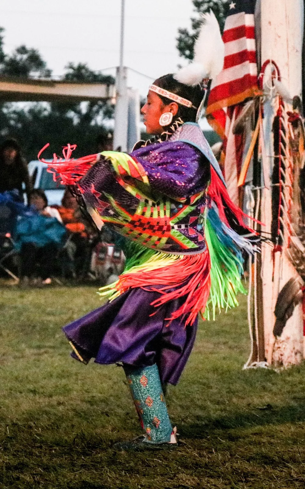 A young woman fancy shawl dancer in purple with bright red, green and yellow fringe spins near a flag-draped pole.