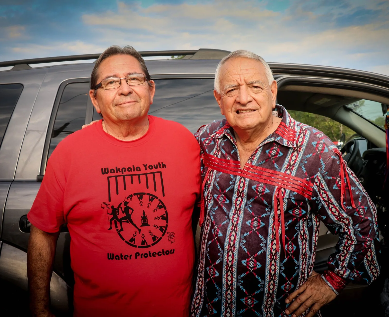 Two elders stand together by an SUV, one in a red Wakpala Youth Water Protectors T-shirt, the other in a patterned ribbon shirt.