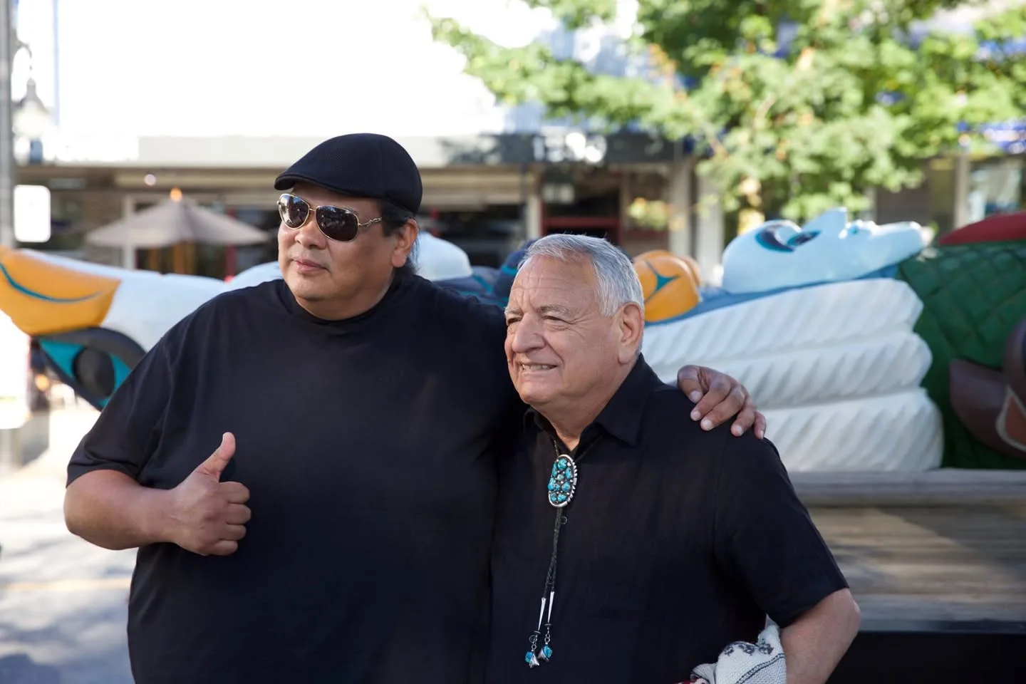 Two men in black shirts stand arm in arm outdoors; one in a cap and sunglasses gives a thumbs up, the other has white hair and a turquoise bolo tie. A carved, painted totem pole lies behind them.