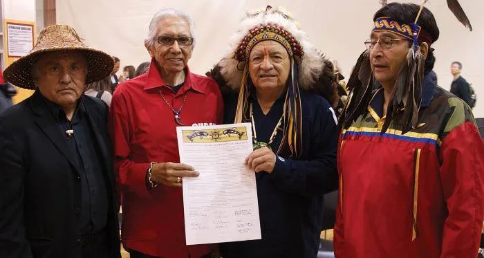 Four Indigenous elders stand together, one in a woven cedar hat, one in a red shirt, one in a feather headdress and one in a beaded headband, holding up a signed treaty document.