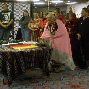 An elder wrapped in a pink blanket leans over a table draped in striped cloth with ceremonial items, as a group of people stands watching behind.
