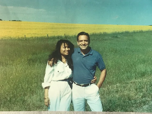 An older faded photo of a smiling couple, Phil and his wife Suthida, standing arm in arm on green grass in front of a bright yellow field under a blue sky.