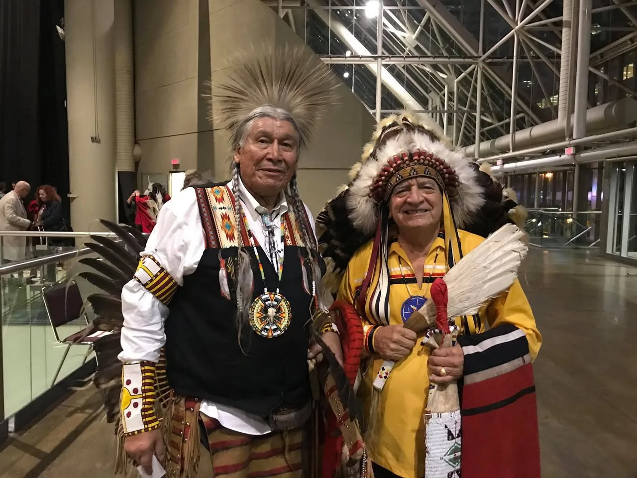 Two elders in full regalia standing together in a large glass-walled hall: one in a porcupine-hair roach and beaded vest, the other in an eagle-feather headdress and yellow shirt holding feathers.