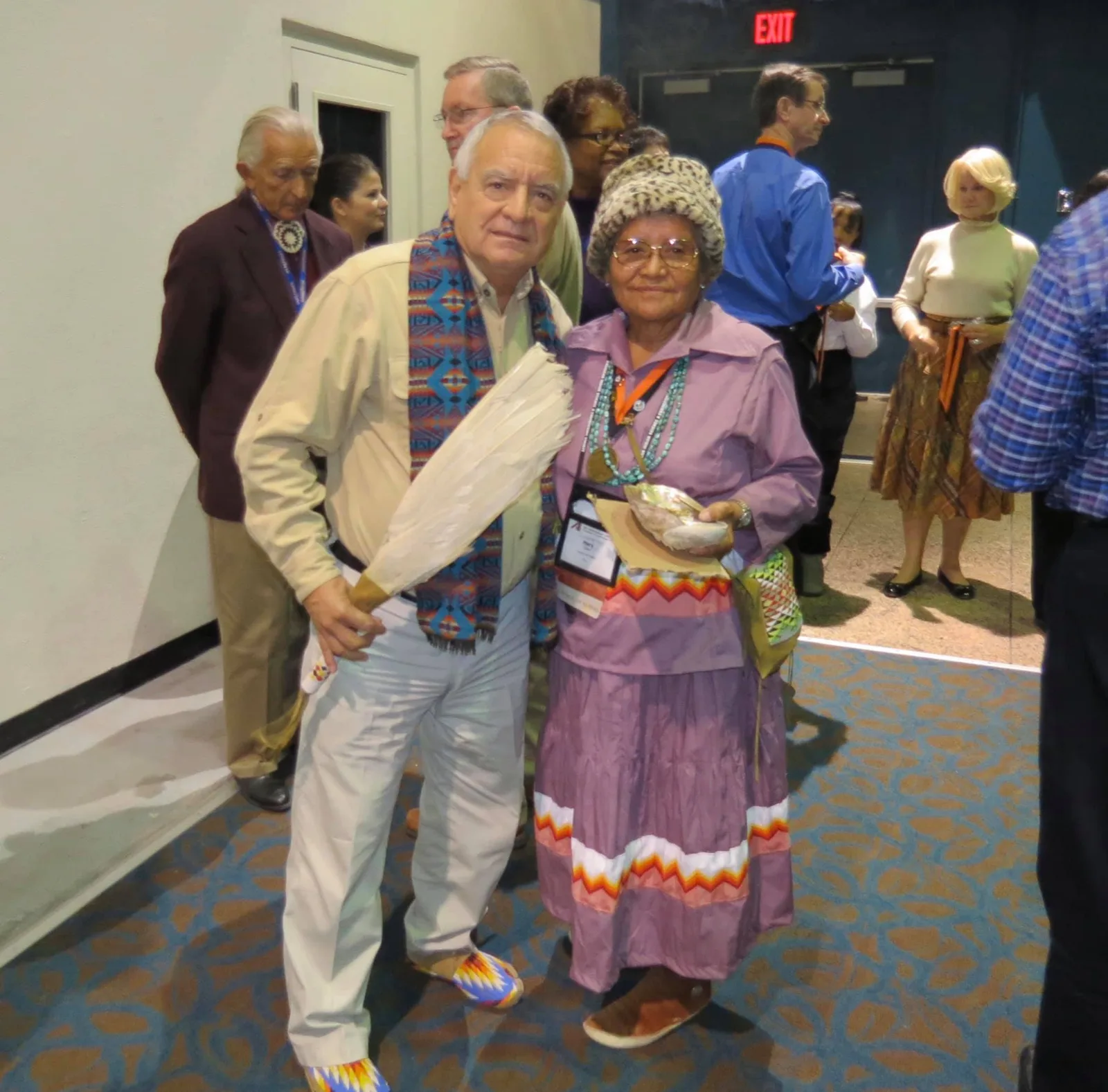 An older man with white hair, holding a white feather fan and wearing a patterned blue scarf and beaded moccasins, stands beside an elder woman in a lavender dress with zigzag ribbon trim, turquoise necklaces and a spotted fur hat, in a crowded conference hallway.