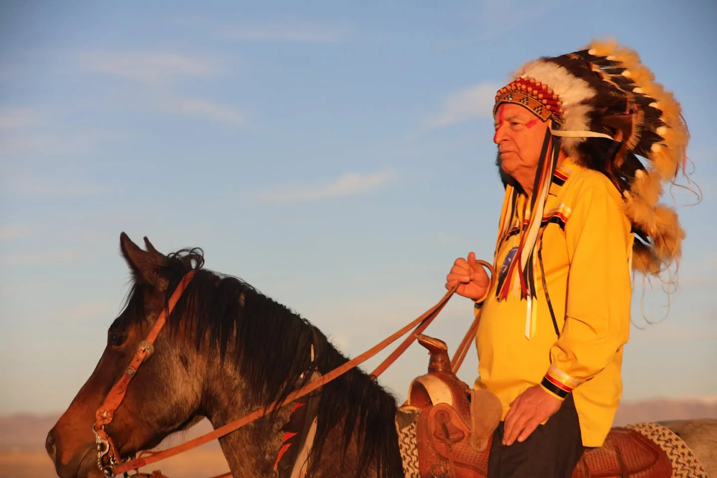 An elder in a large feather headdress, red face paint and a yellow ribbon shirt sits on a brown horse, holding the reins against a clear blue sky at golden hour.