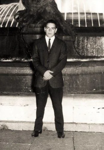 Black-and-white photo of Phil Lane Jr. at 18, a young man in a dark suit and tie standing with hands clasped before the Trevi Fountain in Rome at night.