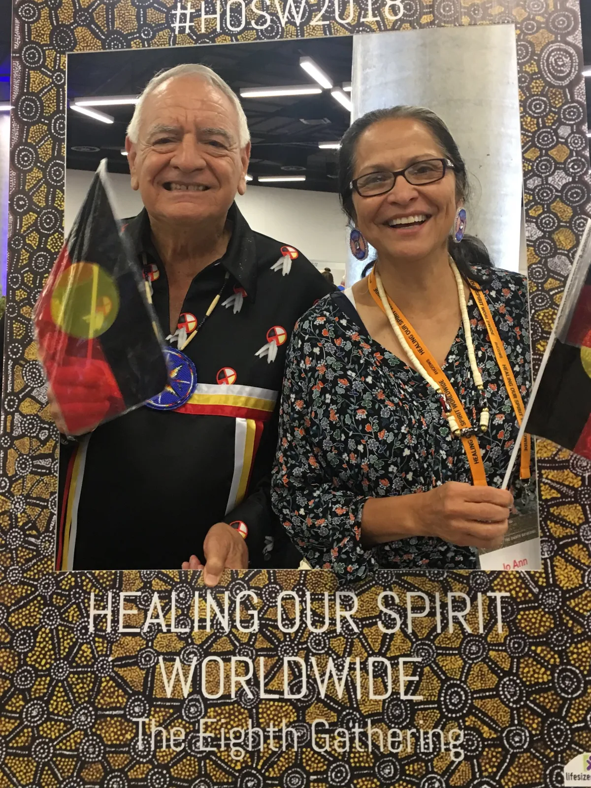 Phil Lane Jr., in a black ribbon shirt with medicine wheel designs, and Jo Ann Kauffman smile holding Aboriginal flags inside a Healing Our Spirit Worldwide Eighth Gathering photo frame.
