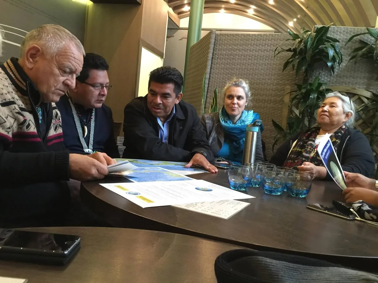 Five people seated around a dark table studying a map of marine sanctuaries and printed documents, with glasses of water and plants nearby.