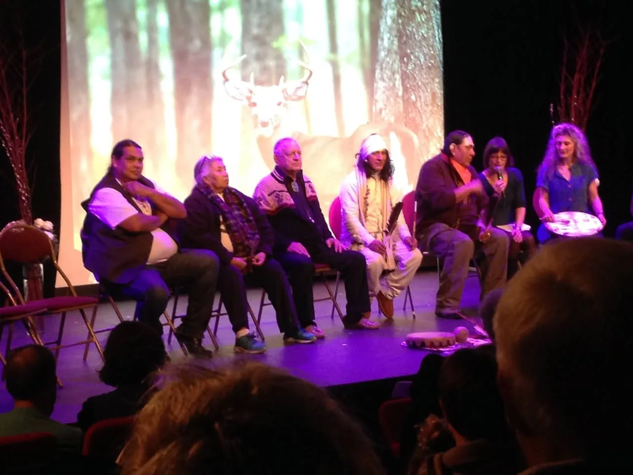 Indigenous elders and guests sit in a row of chairs on a stage before an audience, a projected image of a deer in a forest behind them; a woman speaks into a microphone.