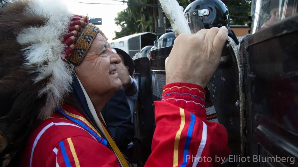 An elder in a feather headdress and red ribbon shirt smiles while holding an eagle feather against the riot shields of helmeted police in a street.