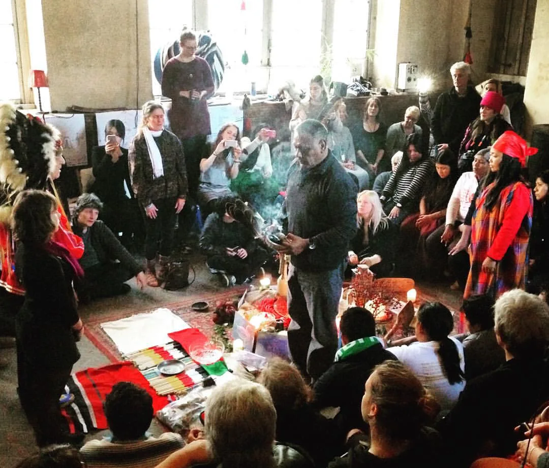 An elder holding a smoking smudge bowl and feather stands at the center of a ceremony, surrounded by a seated and standing crowd, with an altar of candles, flowers and cloths on the floor.