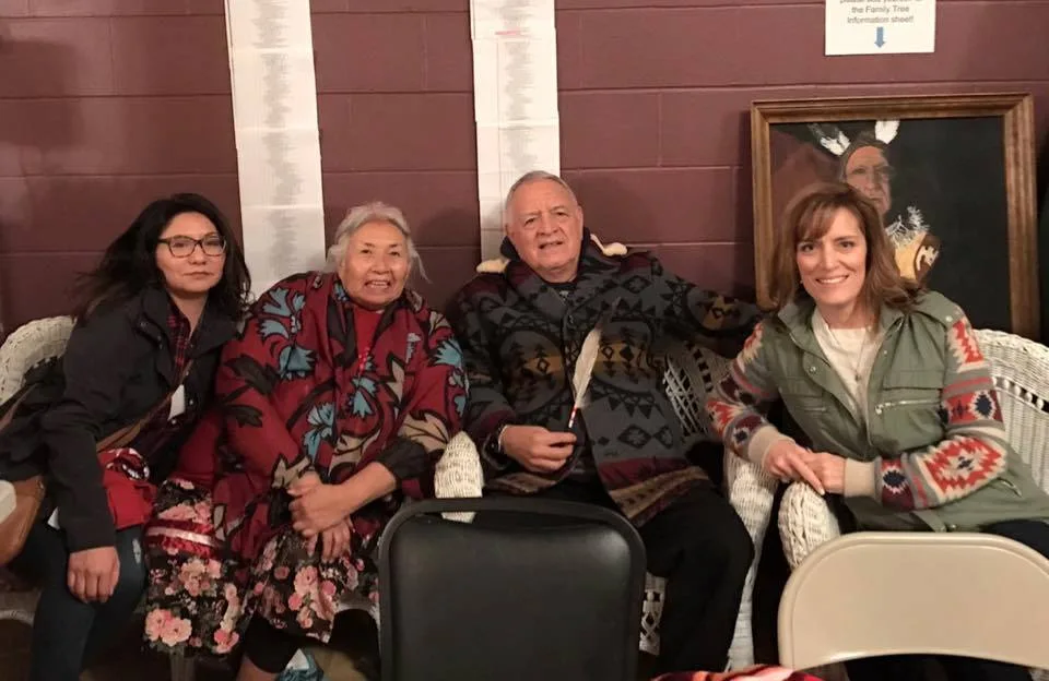 Four people seated on wicker furniture against a maroon wall: a younger woman in glasses, Tankshi Faith in a floral shawl, Phil Lane Jr. holding a feather, and a smiling woman.