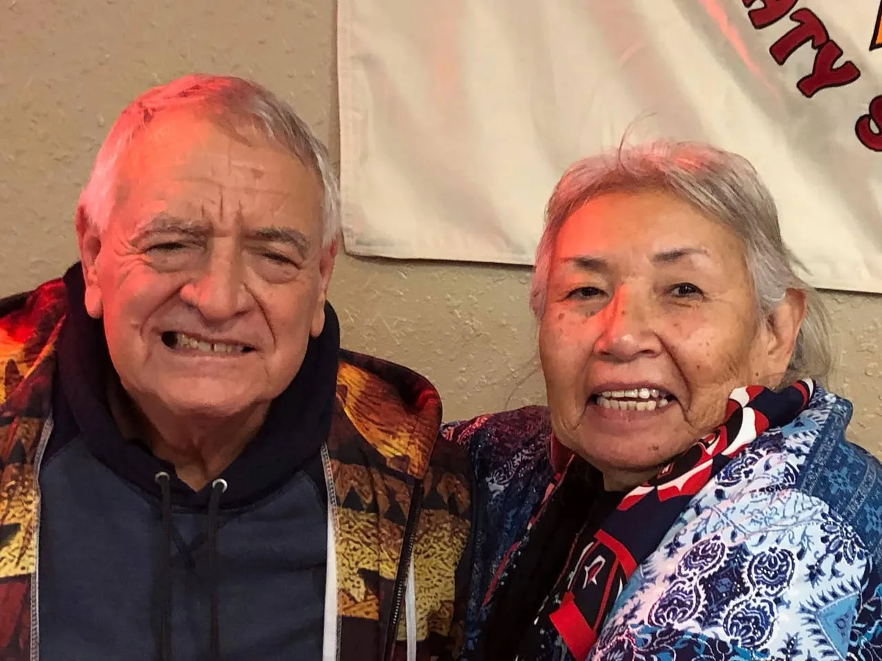Phil Lane Jr. and Tankshi Faith, both with grey hair, smiling close together in colourful patterned jackets in front of a white banner.