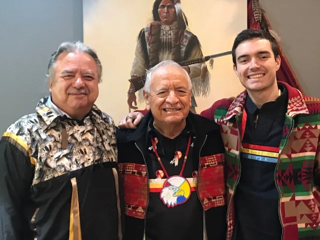 Three men stand smiling together in front of a painting of a Plains warrior: Brother Michael, Phil Lane Jr. in an eagle medallion, and Grandson Hudson in a patterned ribbon shirt.