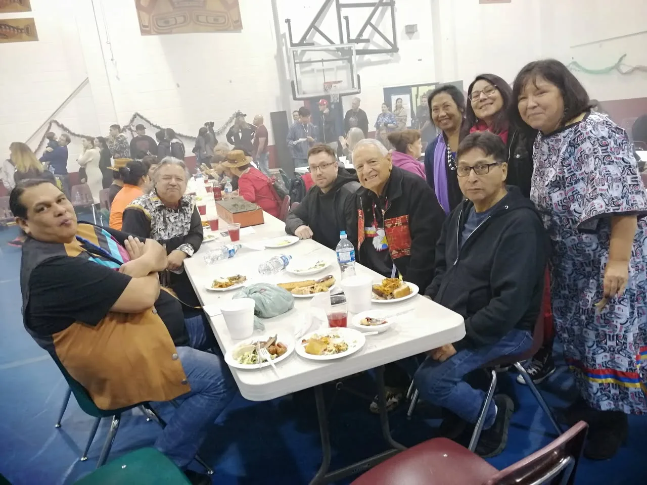 Group seated at a long table with plates of food in a gymnasium hazy with sage smoke; an elder in a red-and-black ribbon vest sits at center, with women in Coast Salish-style dress standing behind.