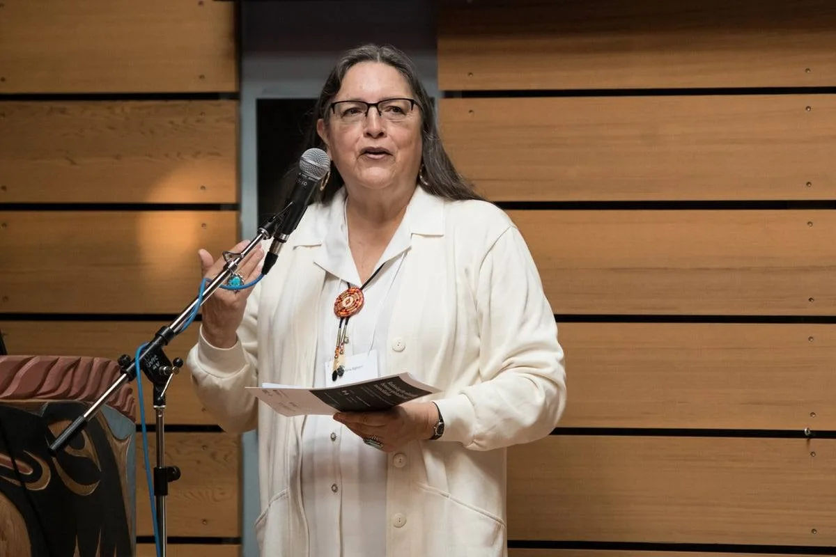 Deloria Bighorn, in a white jacket, glasses and beaded bolo, speaks into a microphone holding a program, before a wooden wall beside a carved Northwest Coast panel.