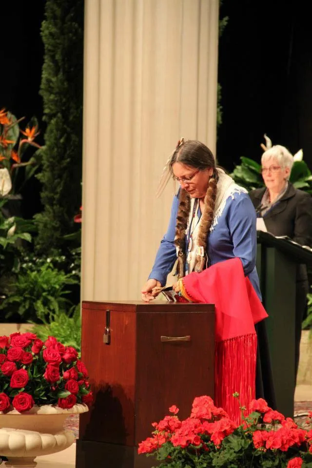 Deloria Bighorn, with fur-wrapped braids and a blue shirt, casts a ballot into a wooden box on a stage with red roses and a white column, as a woman stands at a podium behind.