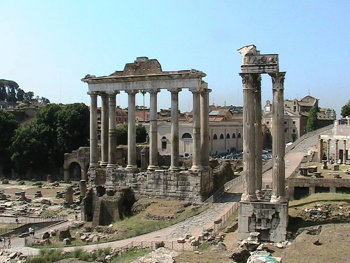 Ruins of the Roman Forum under a clear sky, with the tall columns of the Temple of Saturn and three standing Corinthian columns beside a stone path.