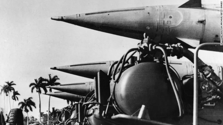 Black-and-white photograph of a row of missiles on launch vehicles, their pointed nose cones aimed upward, with palm trees in the background.