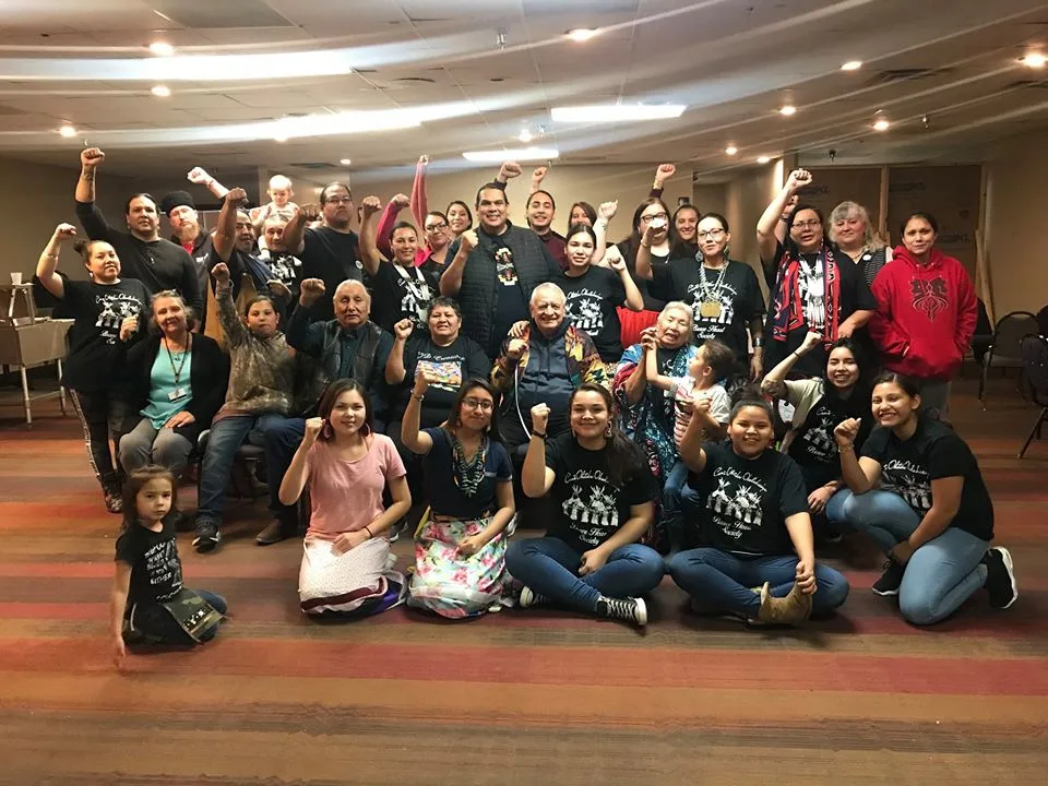 A large group of Indigenous people of all ages pose together in a carpeted hall, many raising fists, several wearing matching black society T-shirts, with elders seated in the center.