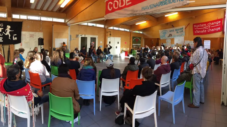 A large group of people seated in a circle of colorful plastic chairs in a wooden hall, listening to speakers; banners include one reading "system change not climate change."