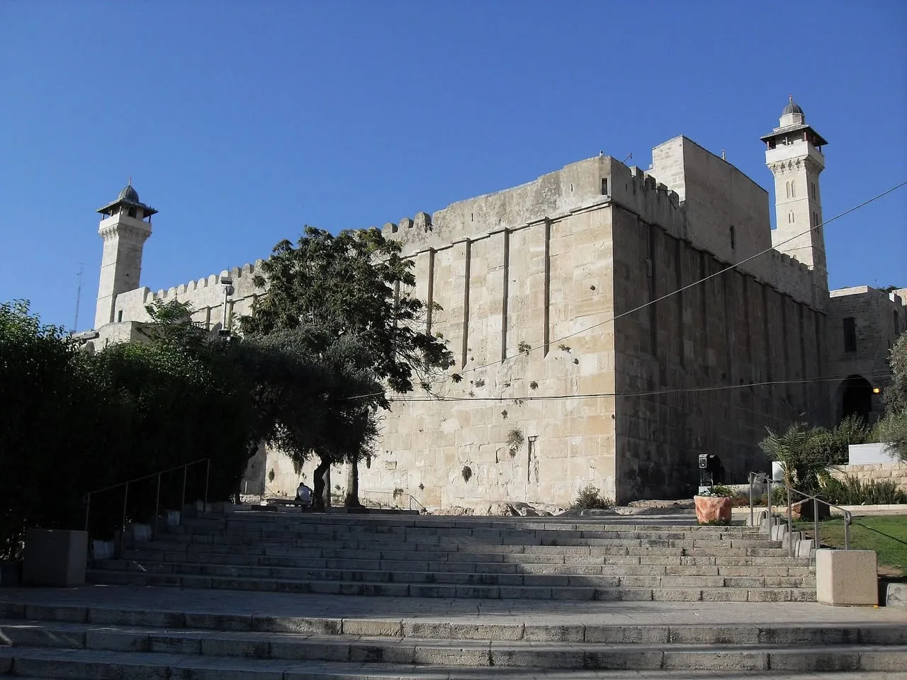 A massive ancient stone building with two minarets under a clear blue sky, approached by wide stone steps, the Cave of the Patriarchs and Matriarchs in Hebron.