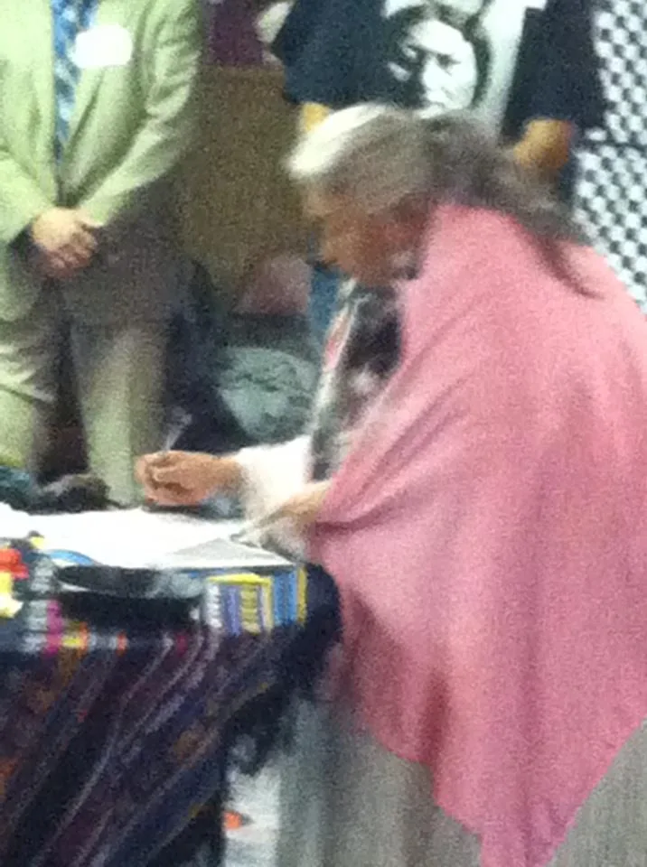 Faith Spotted Eagle, an elder with long gray hair and a pink shawl, leans over a table draped in woven cloth to sign a document as others stand nearby.