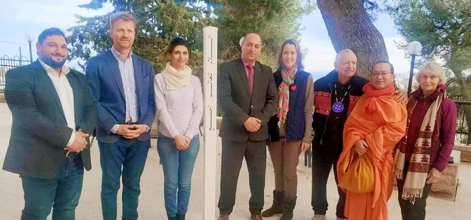 Eight people stand outdoors beside a white peace pole inscribed in several languages, among them a man in a dark suit, a woman in orange robes and an elder in a beaded vest.