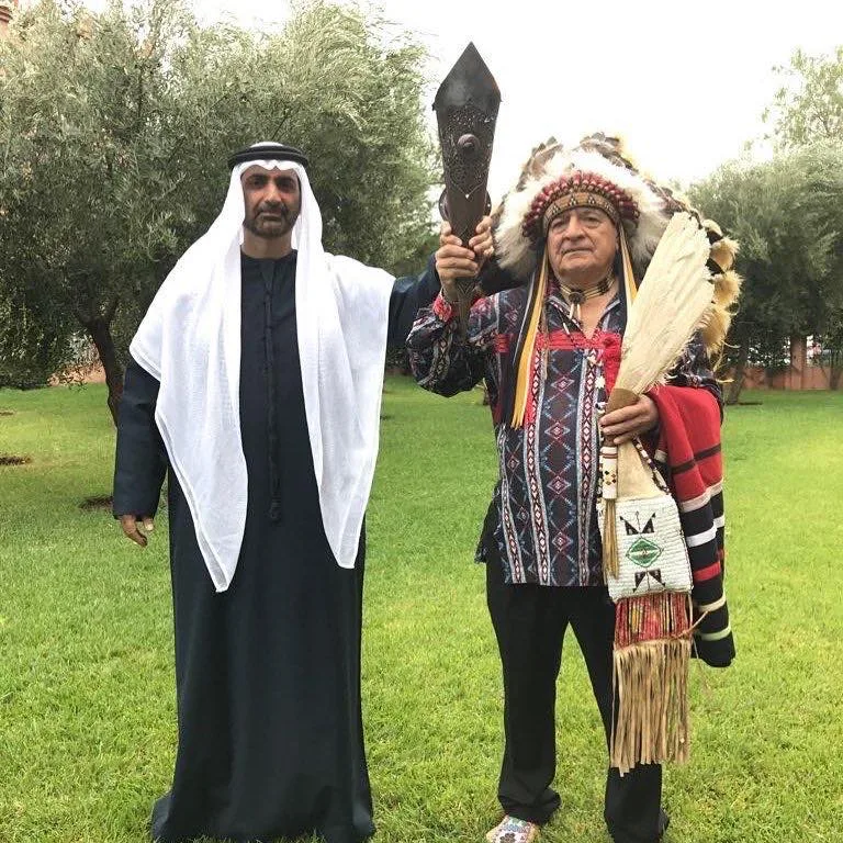 Sheikh Abdul Aziz Al Nuaimi in white headdress and dark robe stands beside an elder in a feather headdress holding a ceremonial staff, on a lawn among olive trees.