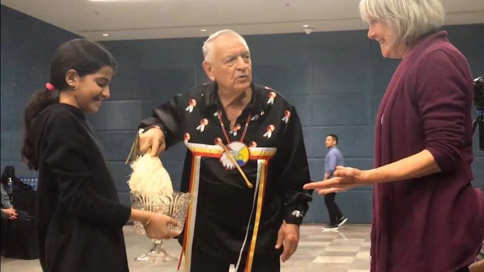 An elder in a black ribbon shirt holds a white feather over a crystal bowl carried by a smiling girl, as a gray-haired woman gestures toward them.