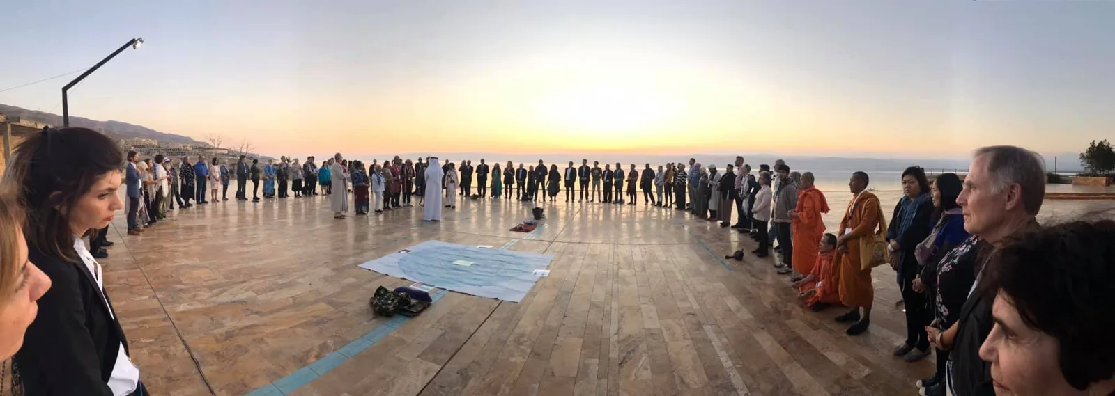 Panorama of a large circle of people of many traditions, including monks in orange robes, standing on a stone terrace at sunrise by the sea.