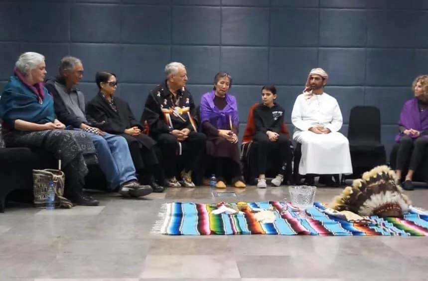 People seated in a circle indoors, including an elder in a ribbon shirt, a girl and a man in white, around a striped blanket holding a feather headdress and a crystal bowl.