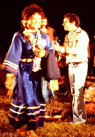 A smiling woman in a blue ribbon dress and beaded necklaces dances at night as a man in a patterned shirt stands behind her.