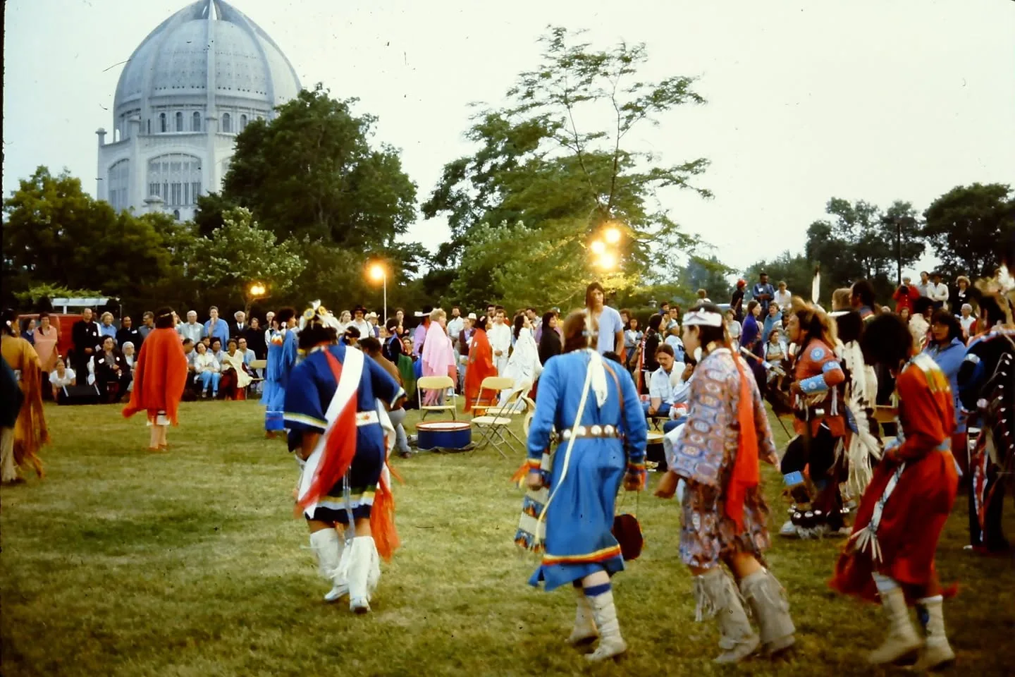 Dancers in colourful regalia move across a lawn at dusk, watched by a large crowd, with a white domed temple rising behind the trees.