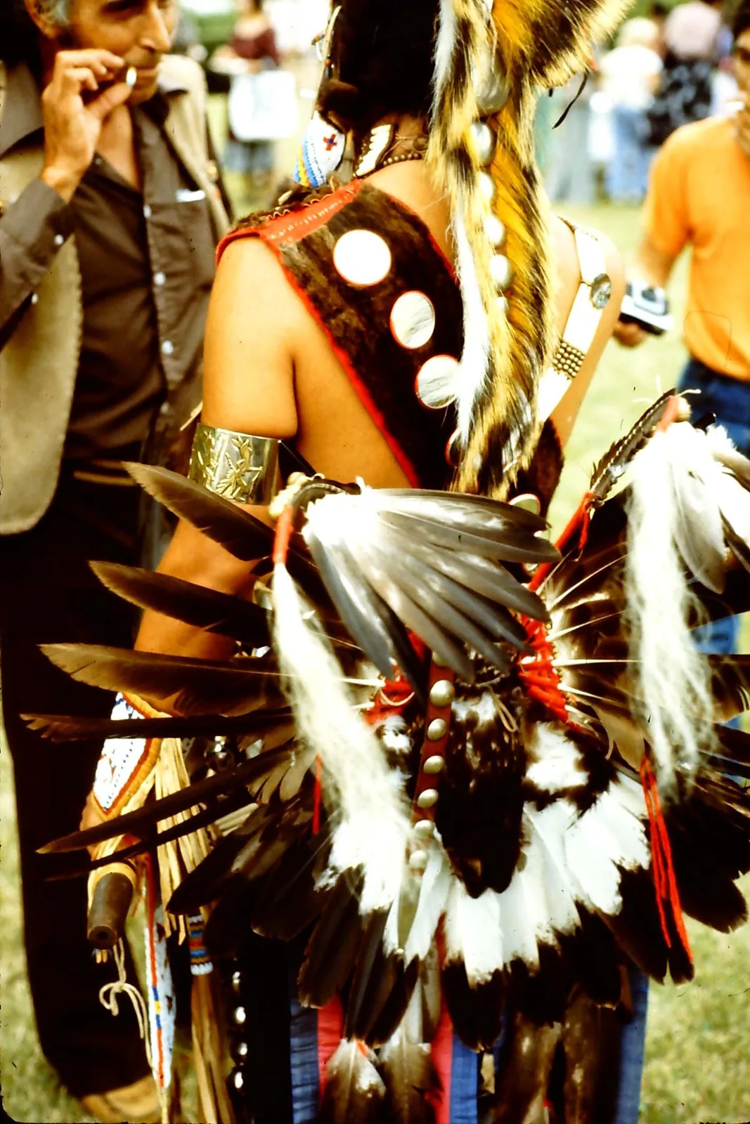 The back of a dancer wearing a feather bustle, roach headdress and vest with round mirrors, beside a man in a brown shirt.