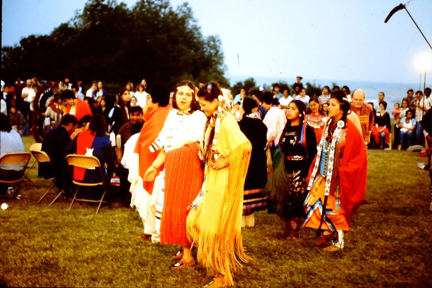 Women in shawls, fringed buckskin and ribbon dresses dance on a lawn at dusk, with a crowd and a lake behind.