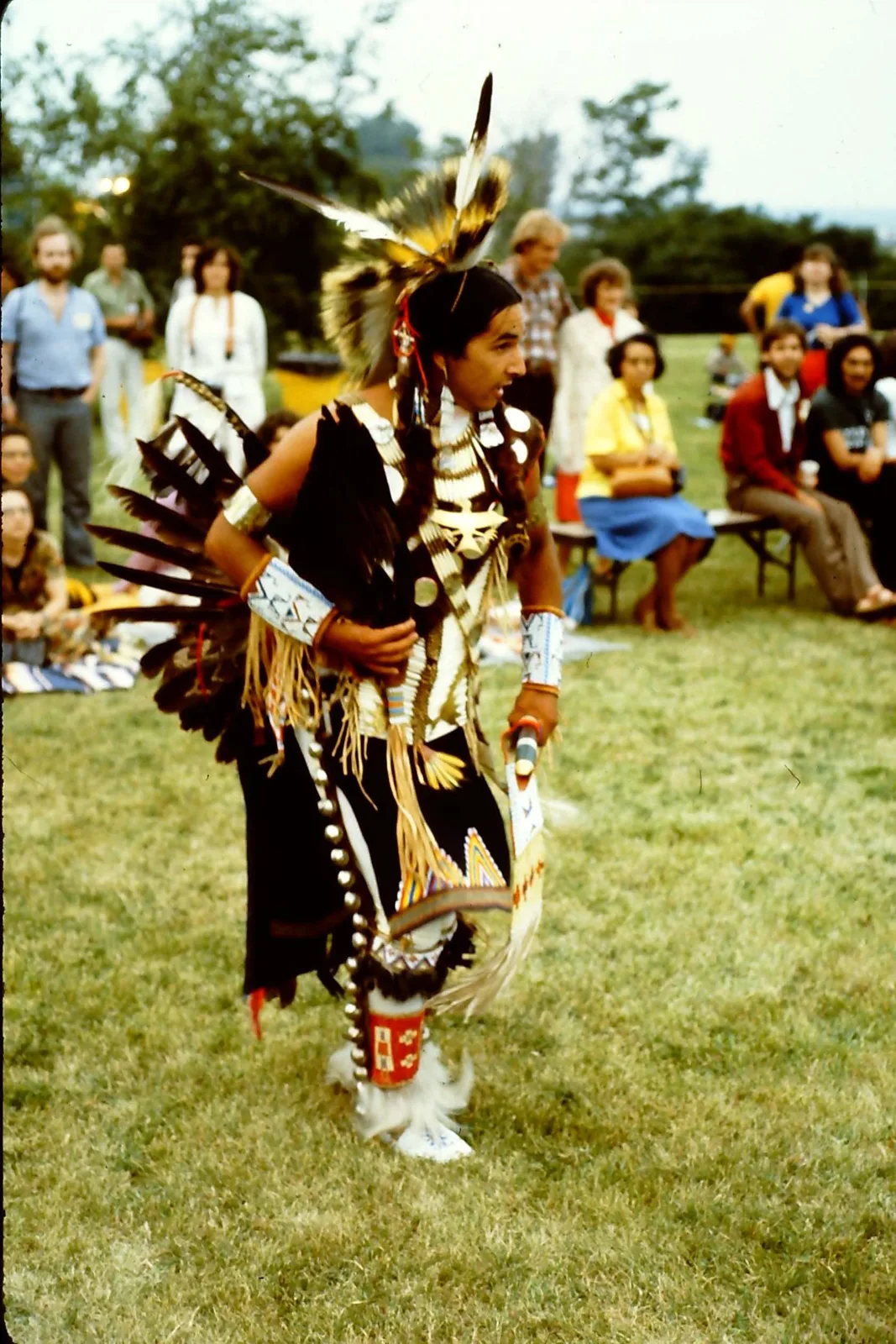 A male dancer in a feathered roach headdress, feather bustle and sleigh bells dances on grass as spectators look on.