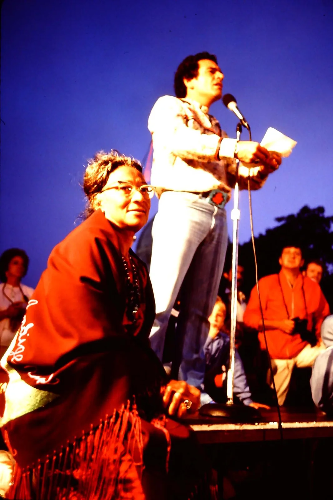 A man speaks into a microphone at night while an elder woman in a red fringed shawl sits in front of him.