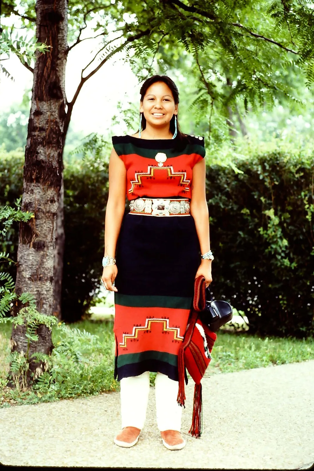 A smiling young woman in a red and black woven dress with a silver concho belt stands beneath a tree.