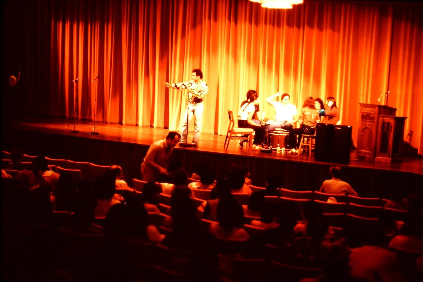 A man speaks at a microphone on a stage with orange curtains while drummers sit nearby and an audience watches.