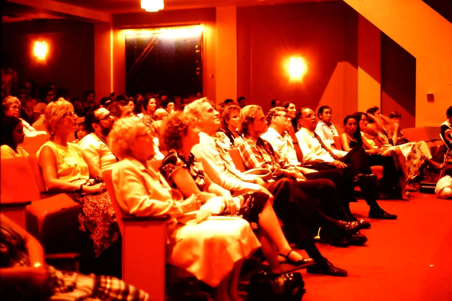 An audience sits in rows in an auditorium bathed in orange light, looking toward the stage.