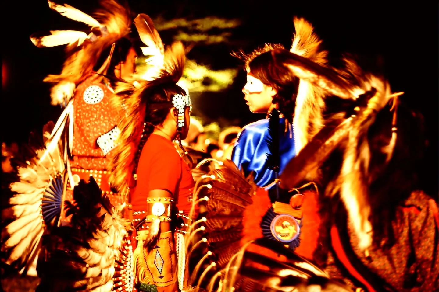 Young dancers in feathered regalia, beadwork and face paint stand close together at night.