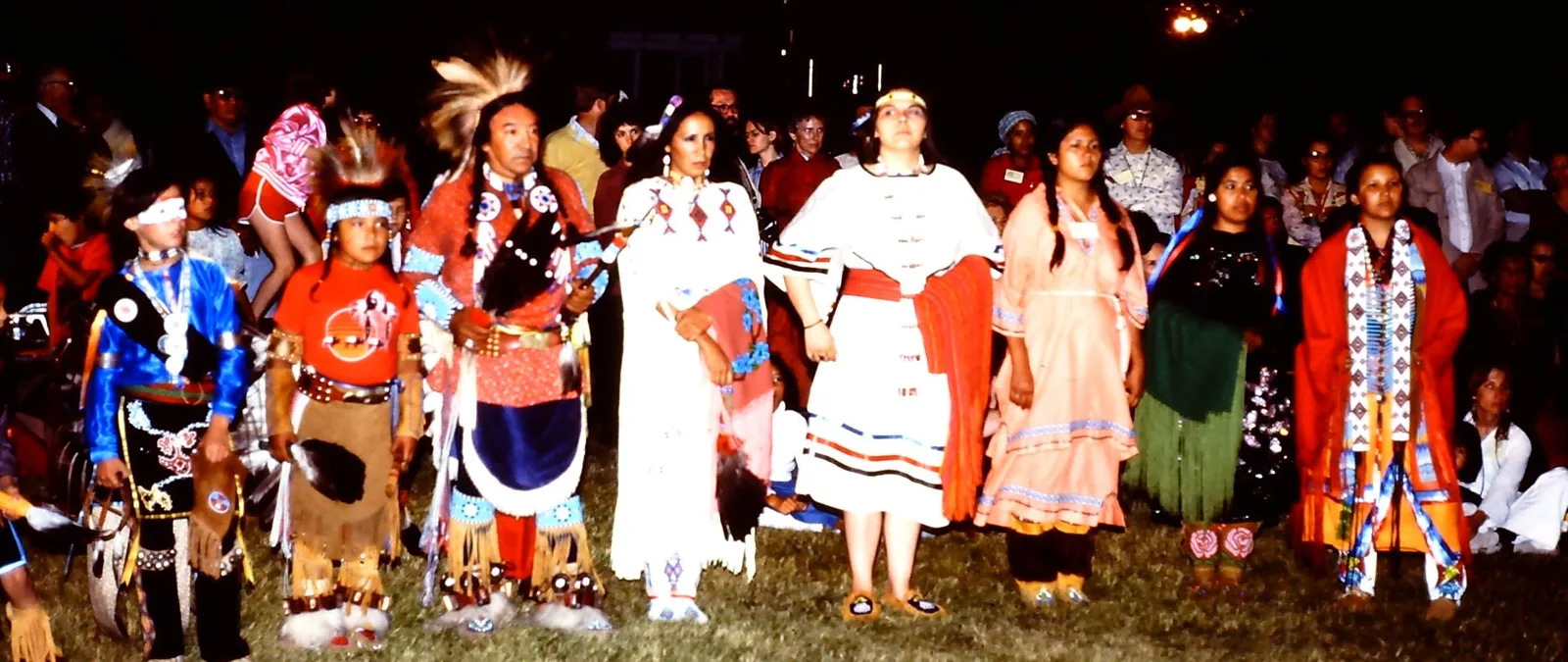 A line of people in regalia, from children to adults, stands side by side at night before a crowd.