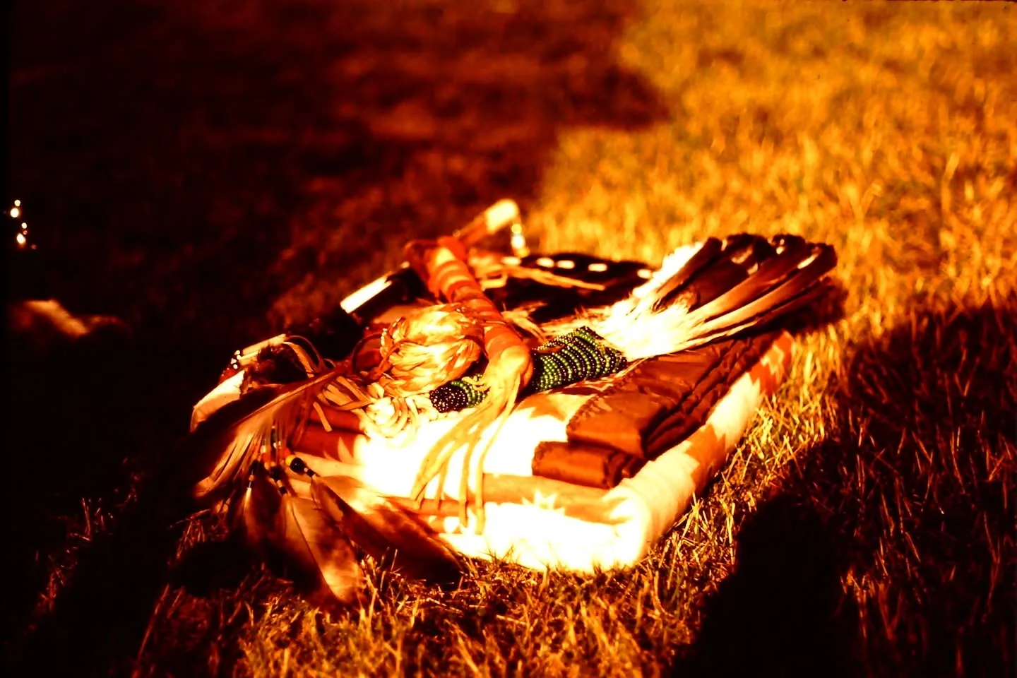 A bundle of feathers and beadwork rests on a folded patterned blanket on the grass at night.