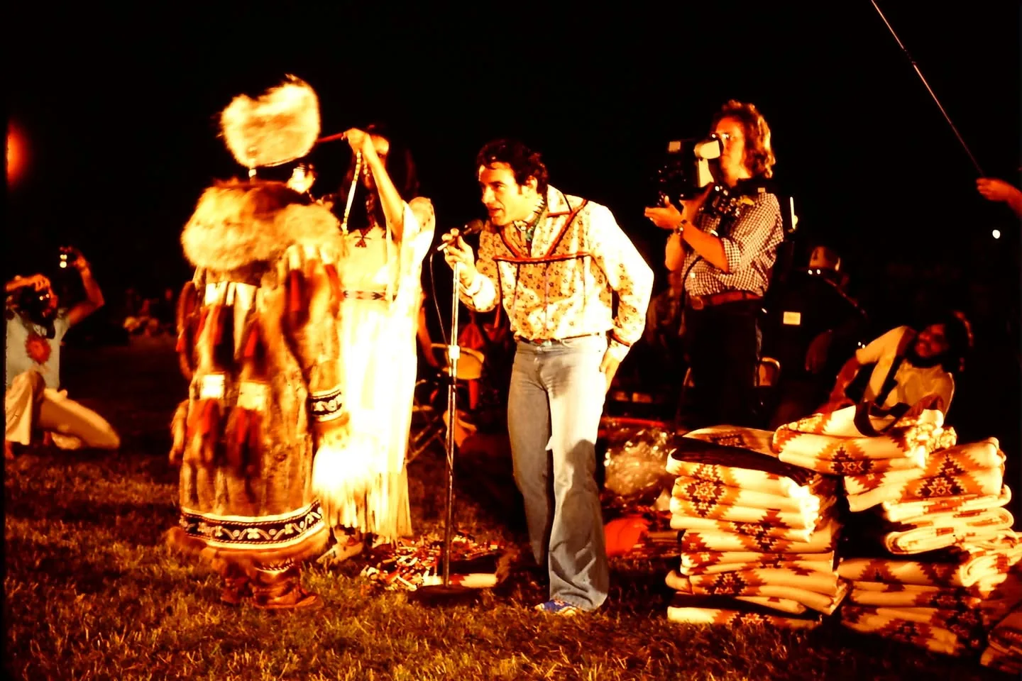 At night, a man speaks at a microphone as a woman places a necklace on a person in a fur parka, beside stacks of blankets.